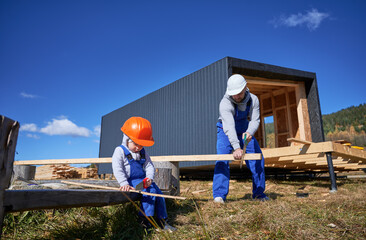 Father with toddler son building wooden frame house. Boy helping his daddy, using hand saw to cut boards on construction site, wearing helmet, blue overalls on sunny day. Carpentry and family concept.