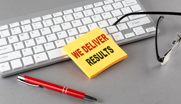 WE DELIVER RESULTS text on a sticky with keyboard, pen glasses on grey background