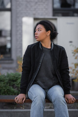 multiracial woman with apathy looking away while sitting on bench on urban street.