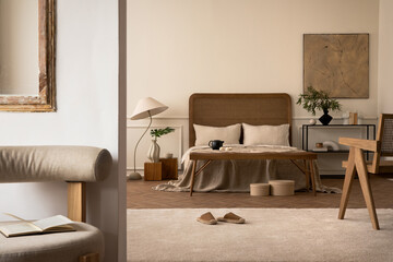 Cozy composition of bedroom interior with mock up poster frame, stylish bed, beige bedding, rattan armchair, bright rug, round box, slippers, lamp and personal accessories. Home decor. Template.