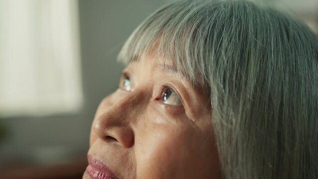 Closeup of lonely Asian wrinkled old lady looking up absent-minded. Elderly woman sit on couch at home living room thinking of life in the past and future missing son and daughter or long lost friends