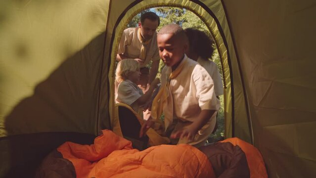Little African American boy scout putting sleeping bag in tent while camping in summer forest with troop and adult scout leader