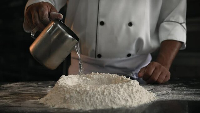 Professional male chef pouring water on flour to make bread dough, closeup shot