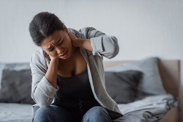 Tensed multiracial woman touching neck while sitting on bed at home.