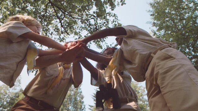 Low angle shot of mid adult Caucasian male scout leader and multiethnic cub scouts putting hands together outdoors in forest on sunny day