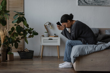 Side view of sad multiracial woman holding smartphone while sitting on bed at home.