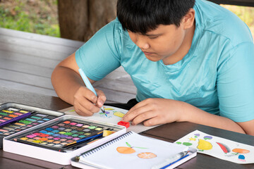 Asian chubby boy in casual blue clothes is practicing drawing and painting by using small watercolor rectangle bars in a paint box on a table on terrace of his summer vacation.