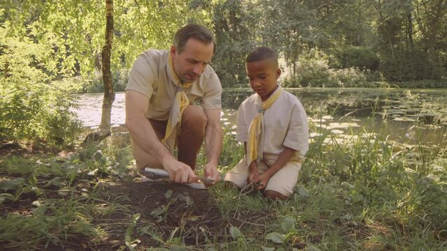 Mid adult Caucasian male scout leader teaching little African American boy scout to make fire with tree branch while camping in forest at summertime