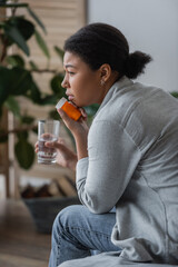 Fototapeta premium Side view of sad multiracial woman holding pills and water while sitting on bed at home.