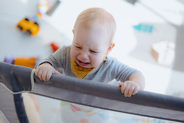 A boy toddler crying in a playpen. Tearful child standing in playpen. baby looking from playpen...