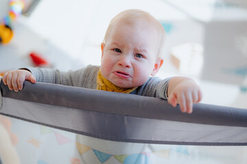 A boy toddler crying in a playpen. Tearful child standing in playpen. baby looking from playpen...