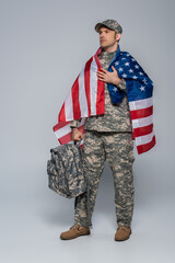 patriotic army soldier in camouflage uniform wrapped in flag of United States of America standing with backpack on grey.