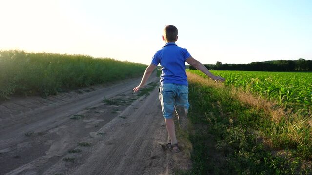 Unrecognizable small boy in sandals running on ground road leaving dust traces. Camera follow for happy child jogging through meadow and rejoicing summertime. Childhood or summer concept. Slow motion