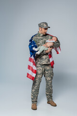 patriotic serviceman in uniform and cap standing with American flag and holding baby boy in arms on grey.