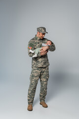 full length of cheerful serviceman in uniform and cap holding newborn son on grey.