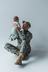 soldier in uniform holding in arms newborn boy while sitting on grey.