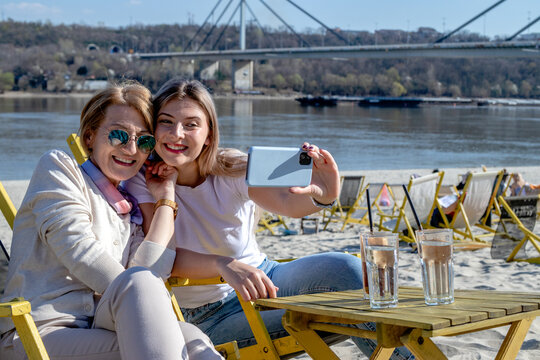 Mother and daughter spend quality time together, bonding, drinking coffee, and taking selfies on the beach bar by the riverbank.