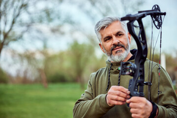 A senior bearded man set up a bow and arrow for shooting, standing in archery.