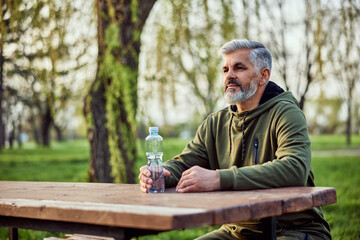 A senior man sitting on a bench in nature holding a bottle of water and looking around.
