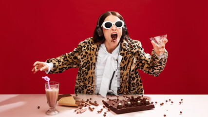 Emotional, eccentric woman in sunglasses and animal print coat, dj on party, playing music and eating chocolate against red background. Concept of pop art, creativity, food, inspiration, fashion