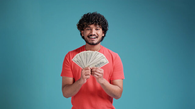 Smiling cheerful happy young bearded Indian man 20s years old wears coral shirt holding showing fan of cash money in dollar banknotes looking camera isolated on plain blue background studio portrait