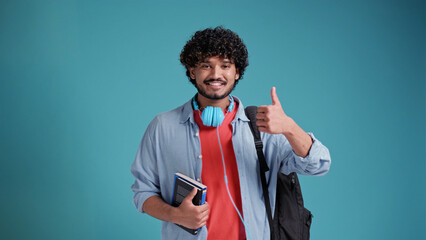 Smiling happy young adult arab or indian man student standing isolated on blue background, showing thumbs up like hand gesture giving recommendation advertising good service or result.