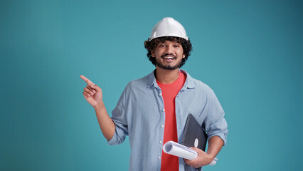 Young indian or arabic architect man with helmet and holding blueprints and laptop over isolated background pointing finger to the side and presenting a product
