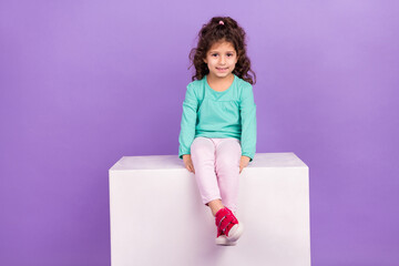 Photo of pretty shiny small kid wear turquoise shirt sitting white platform empty space isolated purple color background