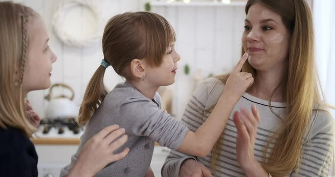 Slow motion shot of playful daughters applying flour on young mother's face while making cookies in kitchen during weekend
