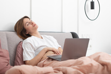 Tired woman lying on bed and working on laptop. Yawning girl looking at the screen of notebook in the bedroom