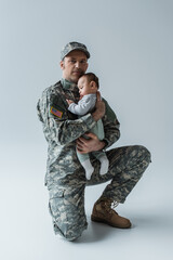 US soldier in uniform holding in arms newborn boy and looking at camera on grey.