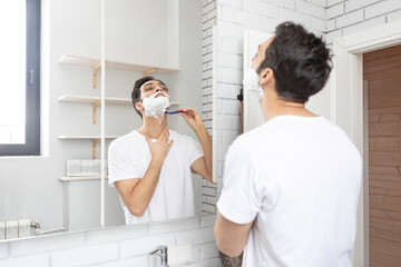 Handsome man looking at the mirror and shaving in the bathroom