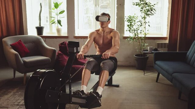Young man with strong, muscular, relief body doing exercises with stationary rowing machine, in VR glasses at home. Online training. Sportive lifestyle, body and health care, fitness, health concept