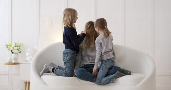Carefree mischievous daughters playing with mother's long hair siting on sofa in living room at home