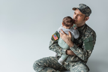 proud father in military uniform holding in arms newborn son while sitting on grey background.