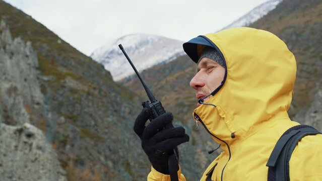 Man in jacket speaks on walkie-talkie standing in mountains. Side view. Search and rescue team worker reports information on portable radioset. Emergency response member use radio communication device