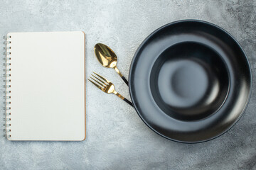 Elegant cutlery set under black dinnerware set and spiral notebook on isolated gray ice background with free space - obrazy, fototapety, plakaty