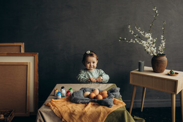 Fototapeta na wymiar Little cute girl with paints and eggs in a studio with beautiful decorations and spring flowers and picture frames