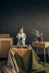 Little cute girl with paints and eggs in a studio with beautiful decorations and spring flowers and picture frames