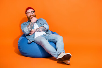 Full length photo of cool dreamy guy dressed denim jacket having rest bean bag looking empty space isolated orange color background