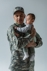 American army soldier in military uniform with cap holding infant son in arms isolated on grey.