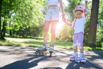 Mother teaching child daughter to skate on inline skates rollers in public park in summer. Family leisure outdoor sport activity game. Copy negative space