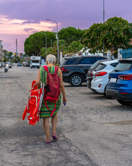 Real lonely woman in beach shoes with a beach chair walking on the road to the parking lot,...