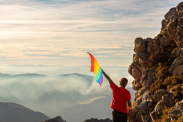 mountaineer man making summit on a mountain and waving a rainbow lgbt pride flag at sunset. lgbt...