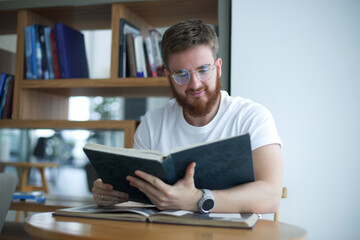 Portrait of young bearded university or college happy student in glasses sitting in library and read book. Education concept.
