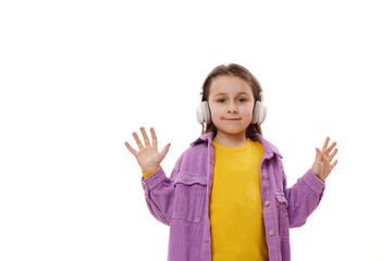 Caucasian happy adorable little girl in bright yellow sweater and purple jacket, wearing wireless headphones, looks and camera and shows her hands palms up, isolated over white background. Copy space