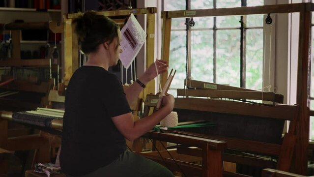 Woman organizes and wraps yarn from spool onto shuttle while sitting at the loom