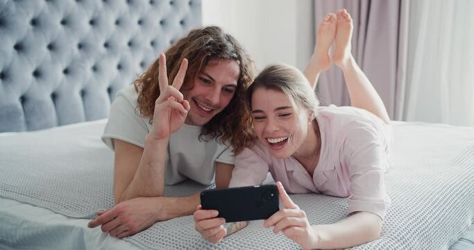 Attractive happy young couple laying on bed in the bedroom, making silly faces, taking a selfie using mobile
