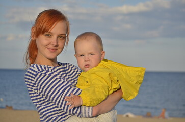 A young mother and a small child in her arms are happy on the background of the sea. Close-up portrait. Mom and baby on the sea, family vacation concept