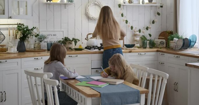 Caucasian young mother cooking food on stove while daughters writing homework on table in kitchen at home